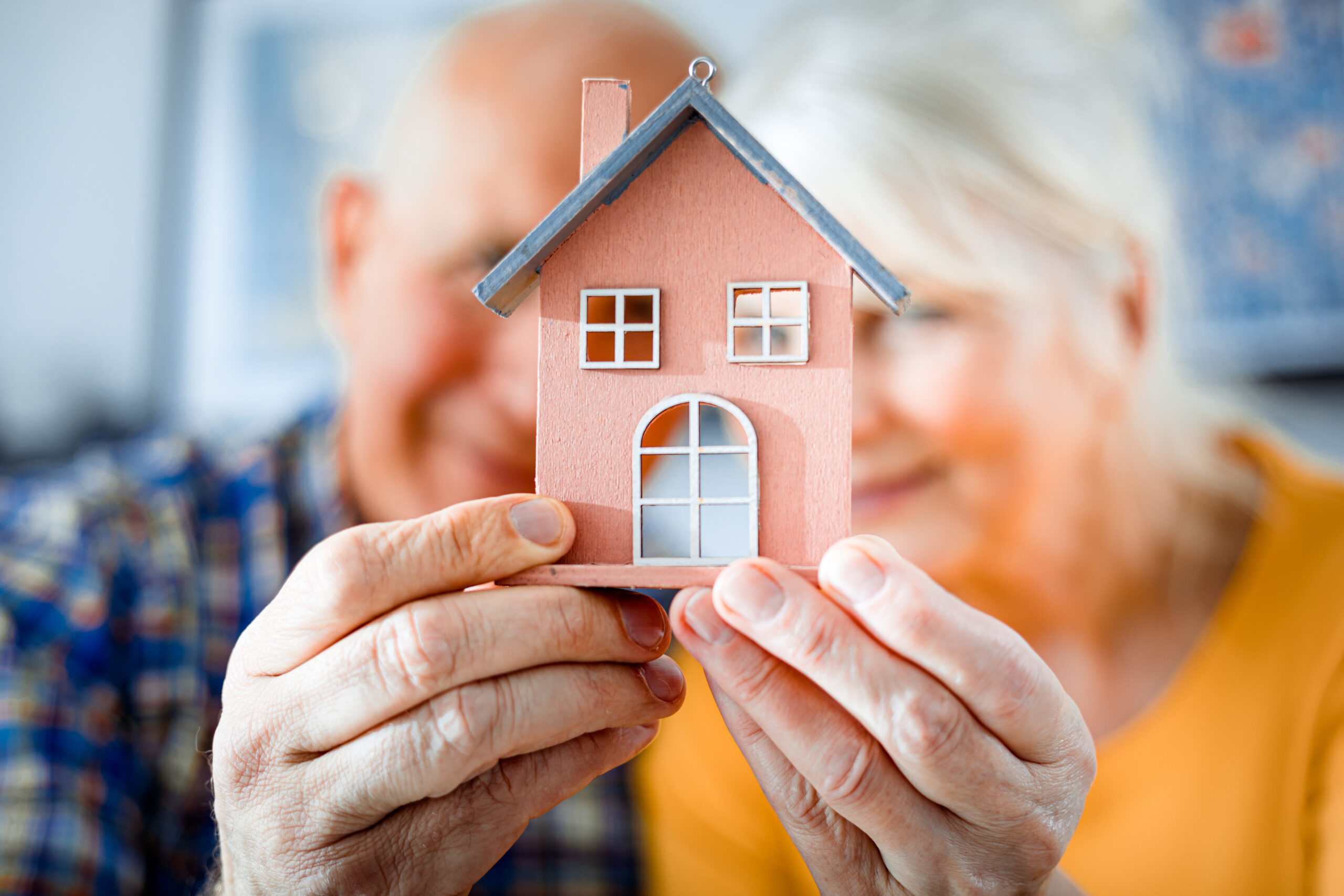 An older couple (blurred in the background) hold a freshly painted 3D model of a home