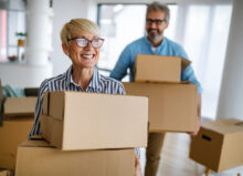 An older couple is standing in a home surrounding by boxes. Both the woman and man are holding two cardboard boxes and smiling.