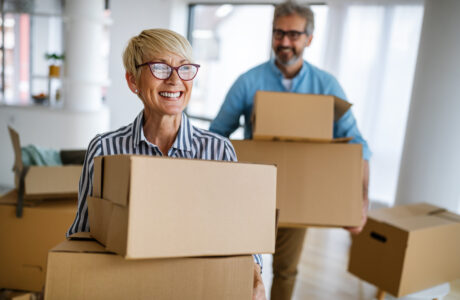 An older couple is standing in a home surrounding by boxes. Both the woman and man are holding two cardboard boxes and smiling.