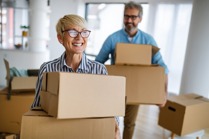 An older couple is standing in a home surrounding by boxes. Both the woman and man are holding two cardboard boxes and smiling.