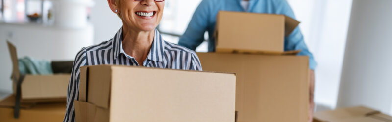 An older couple is standing in a home surrounding by boxes. Both the woman and man are holding two cardboard boxes and smiling.