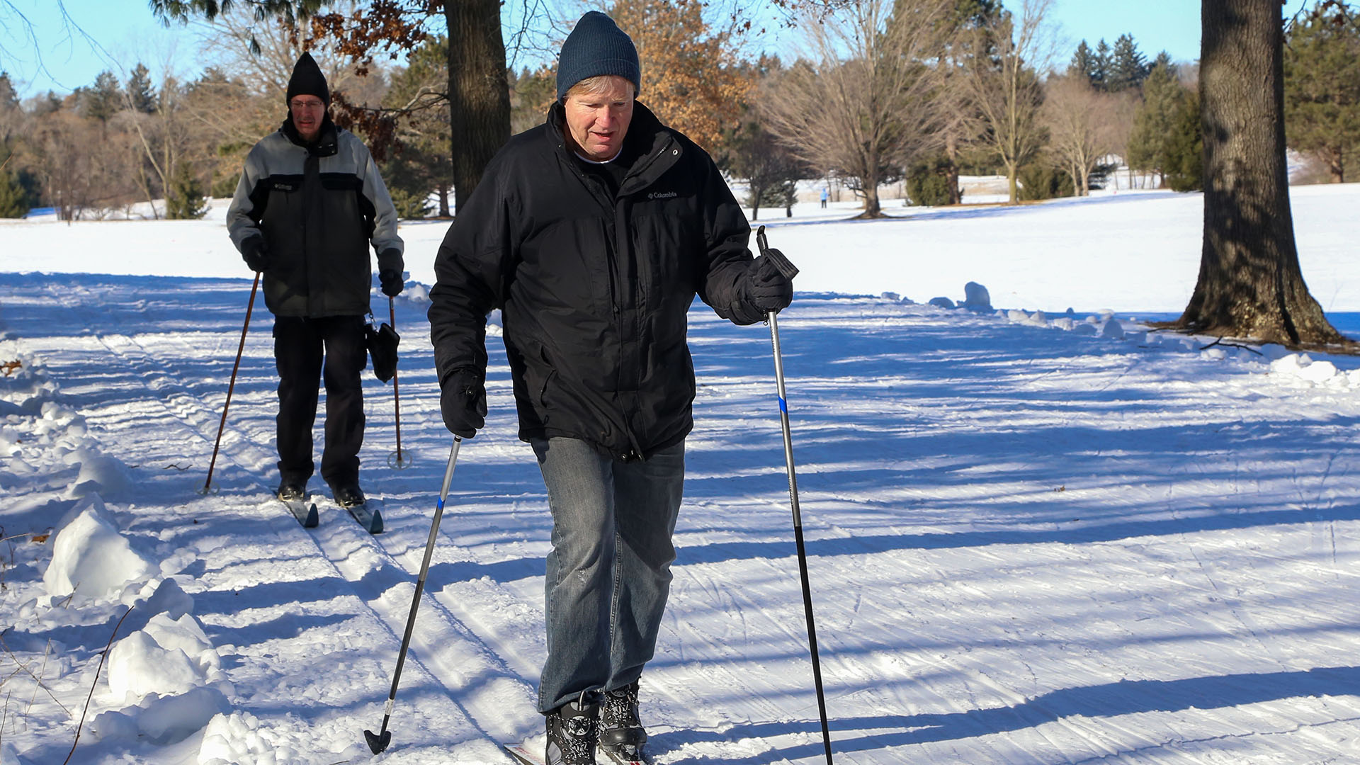Two older men are skiing on a flat, snow-covered trail at Stony Creek Metropark. 